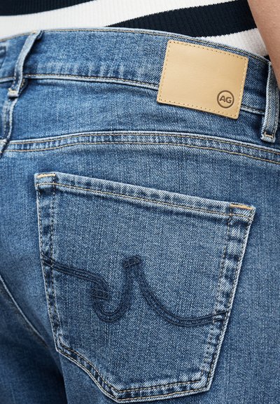 Τζιν παντελόνι από μπλε τζιν με πίσω τσέπη διακοσμημένη με ραμμένο σχέδιο και ταμπελάκι από ταμπά δέρμα με την επωνυμία «AG» στη μέση, φορεμένο με ριγέ τοπ.
