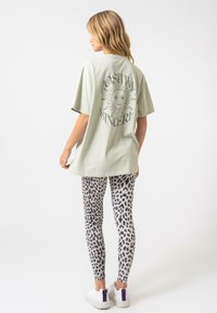 Světlězelené oversized tričko s grafikou slunce a textem na zadní straně, spárované se šedými legíny s leopardím potiskem a bílými teniskami s fialovými akcenty.