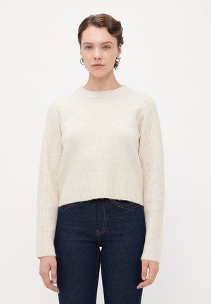 Kvinne med kort hår som har på seg sølv hoop-øreringer, en kremfarget ribbestrikket genser og mørkeblå high-waisted jeans, stående mot en hvit bakgrunn.