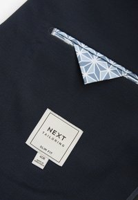 Tejido texturizado en color navy con una etiqueta de corte ajustado. Un pañuelo de bolsillo azul claro con un patrón geométrico asoma del bolsillo del pecho.