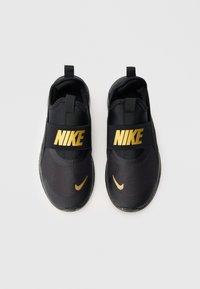 Fekete Nike slip-on sneaker pár arany logókkal és elasztikus pántokkal a tetején, felülnézetből, fehér háttéren.