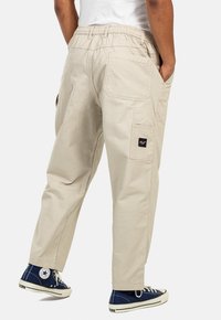 Beige Cargo-Hosen mit elastischem Bund, Seitentaschen und einem Logopatch. Die Beine sind locker geschnitten und mit gesammelten Bündchen versehen. Kombiniert mit blauen Turnschuhen.