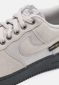 Šedá semišová a síťovaná teniska s černou gumovou podrážkou. Obsahuje logo Nike, šněrování vpředu a štítek Cordura pro odolnost.