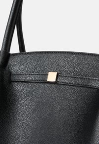 Bolso tote de cuero negro con una superficie texturizada. Presenta un acento cuadrado dorado y dos asas resistentes. Diseño simple y moderno.