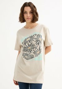 Camiseta de algodón beige con un gran gráfico floral en gris, acentos amarillos y texto turquesa. Corte relajado y mangas cortas.