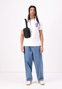 Black crossbody bag with a smooth texture and zip closure, worn by a person in a white t-shirt and blue jeans with a relaxed fit.