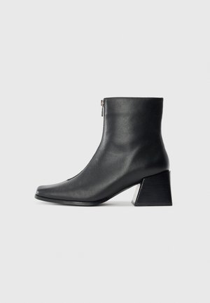 Bota de tornozelo em couro preto com bico quadrado, fecho lateral e salto bloco contra um fundo claro plano.