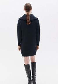 Abito maglione blu navy con colletto ampio, maniche lunghe e polsini e orlo a coste. Abbinato a stivali neri fino al ginocchio. Visione posteriore.