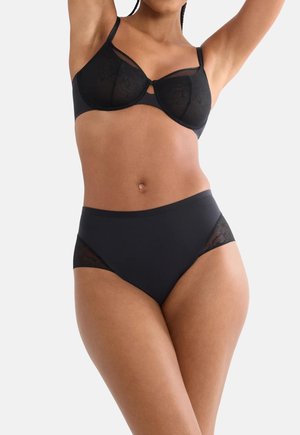 Kobieta w czarnym przejrzystym koronkowym biustonoszu i majtkach z wysokim stanem z koronkowymi panelami, uniesionymi ramionami, na jasnym jednolitym tle.