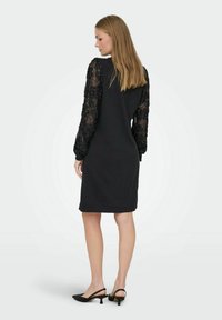 Robe noire avec des manches longues en dentelle, design ajusté, longueur à genoux, texture lisse, accompagnée de talons slingback noirs.