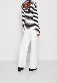 Gestreiftes Langarm-Oberteil mit runder Rückenschneidung und Bindedetail, kombiniert mit weißen weit geschnittenen Hosen und schwarzen Plateauschuhen.