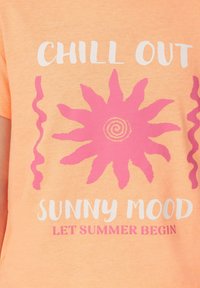 Pfirsichfarbenes T-Shirt mit einem pinken Sonnengrafik mit Wellenmotiven und weißem Text, der "CHILL OUT SUNNY MOOD LET SUMMER BEGIN" liest.