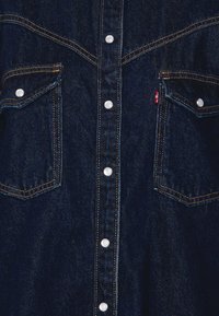 Mörk denimskjorta med dubbla bröstfickor, silverfärgad tryckknapp, kontrasterande gul sömnad och en subtil texturerad yta.