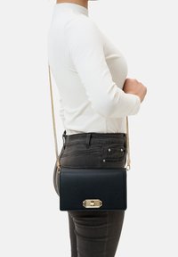 Schwarze Leder-Crossbody-Tasche mit einem goldenen Metallverschluss, rechteckiger Form und einem langen Kettenriemen, die an der Hüfte getragen wird. Glatte Textur.