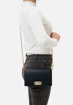 Schwarze Leder-Crossbody-Tasche mit einem goldenen Metallverschluss, rechteckiger Form und einem langen Kettenriemen, die an der Hüfte getragen wird. Glatte Textur.