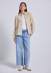Beige corduroy button-up shirt over een witte top, gecombineerd met lichtblauwe wijde jeans en witte sneakers. Eenvoudig ontwerp en neutrale tinten.