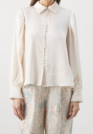 Frau trägt eine cremefarbene, strukturierte Langarmbluse mit stoffbezogenen Knöpfen und hellblaue Hose mit Blumenmuster.