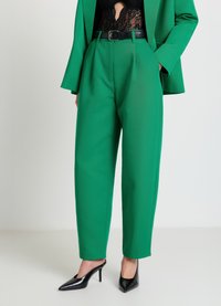 Pantalon vert sur mesure avec une taille haute, des plis et une ceinture noire. Associé à un body en dentelle noire et des escarpins noirs à bouts pointus.