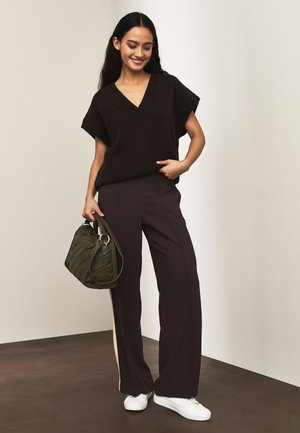 Naeratav naine, kes kannab musta V-kaelusega varrukateta kampsunit, musti laiade säärega pükse küljeribaga, valgeid tossusid ning hoiab oliivrohelise värvi käekotti.