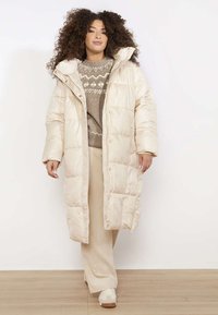 Długa beżowa kurtka puchowa z pikowanym wzorem, kapturem i przednim zamkiem błyskawicznym. Nosi się ją na wzorzysty sweter i beżowe spodnie prążkowane oraz trampki.