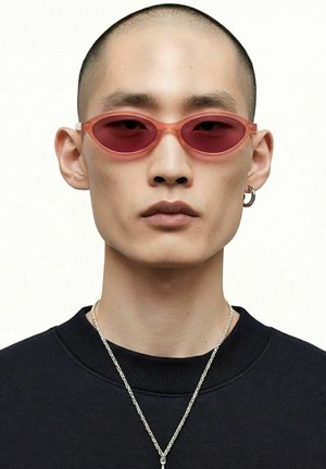 Jeune personne au crâne rasé portant des lunettes de soleil ovales roses, une boucle d'oreille argentée en anneau, un collier chaîne argenté et un t-shirt noir à col rond sur fond blanc.