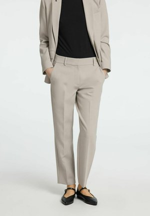 Light beige tailored trousers with a smooth texture and creases, high waistband, and side pockets, paired with black flat shoes.