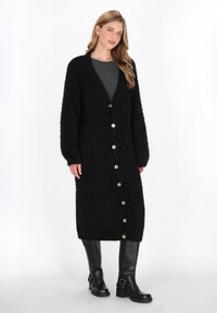 Schwarzer, langer Cardigan aus weichem, flauschigem Material mit einem tiefen V-Ausschnitt, großen goldenen Knöpfen und langen Ärmeln. Kombiniert mit schwarzen kniehohen Stiefeln.