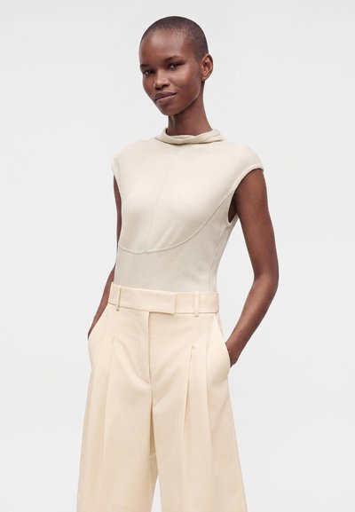 Femme portant un haut beige sans manches et un pantalon large crème taille haute, les mains dans les poches, debout devant un fond clair uni.