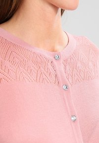 Ljusrosa kofta med ett texturerat stickat mönster på oket, försett med tre klara stenar som knappar och en slät tygfinish.