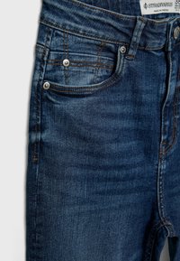 Jeans de mezclilla azul oscuro con un diseño ajustado, que cuentan con herrajes plateados, costuras en contraste y múltiples bolsillos. Tejido texturizado con un ligero brillo.