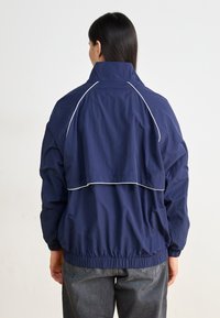 Marineblå vindjakke med hvid piping, elastisk talje og lagdelt rygdesign. Letvægts, åndbart stof.