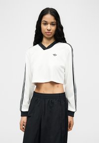 Γυναίκα με μακριά σκουρόχρωμα μαλλιά φοράει λευκό κοντό φούτερ με μαύρες ρίγες και μαύρο παντελόνι, ποζάροντας μπροστά σε απλό ανοιχτόχρωμο φόντο.