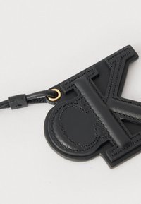 Porte-clés en cuir noir avec lettres brodées en relief "C" et "K", attaché à un cordon noir avec un anneau en métal doré.