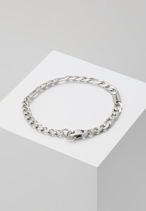 Bracelet chaîne ton argent avec maillons plats imbriqués et fermoir mousqueton, présenté sur un socle géométrique blanc.