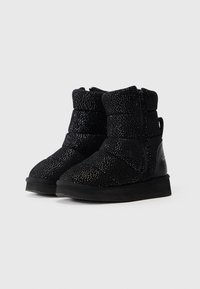 Botas negras de tobillo con textura brillante, diseño almohadillado, cremallera lateral y suelas planas sobre un fondo claro y liso.