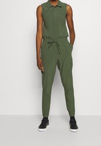 Olivově zelený bezramínkový overal s límečkem, stahovací páskou a zúženými nohavicemi. Nosený s černými teniskami.