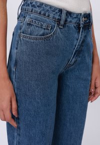 Detail osoby nosící modré džíny z denimu s viditelným předním kapsou, poutky na opasek a zapínáním na knoflík, ruce volně podél těla.