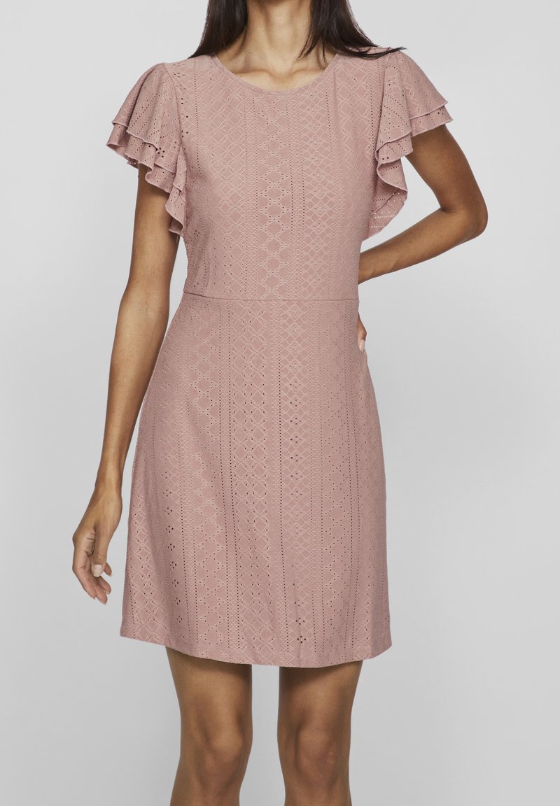 Femme portant une robe rose clair à manches courtes avec des motifs en œillets et des manches à volants superposés, debout devant un fond gris uni.