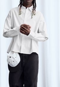 Fehér csíkos, laza szabású ing sötét farmernadrággal párosítva. Egy fehér sapka fekszik a zseben, fekete karimával.