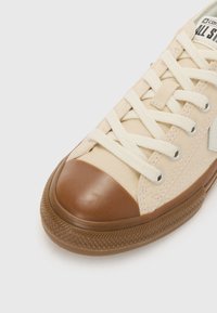 Beige lerretsko med brunt gummitåhette og såle, med kremfargede lisser, metalløyer og logo på tungen.