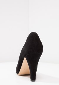 Chaussure noire en daim à talon haut avec un bout pointu et un talon effilé. La semelle est de couleur claire et présente une texture lisse.