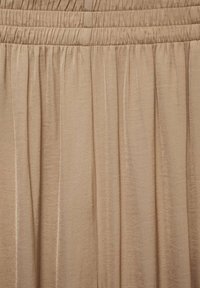 Tissu beige fluide avec une texture lisse, comportant un élastique froncé à la taille et des plis verticaux pour un tombé supplémentaire.