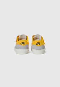 Nike SB-skor med en övre del av gult tyg, grå mockasektioner och en vit gummisula. Inkluderar ett kardborreband och logodetalj på hälen.