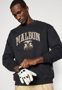 Μαύρη μπλούζα με κουκούλα που έχει το κείμενο "MALBON" και έμβλημα σε χρυσή κεντητική. Το μοντέλο φοράει ένα λευκό γάντι γκολφ με κομμένες τρύπες, αναδεικνύοντας αθλητικό στυλ.