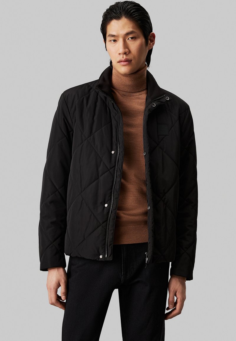 Chaqueta negra acolchada con botones a presión, cuello alto y bolsillos laterales, llevada sobre un suéter de cuello alto marrón. Diseño simple y moderno.