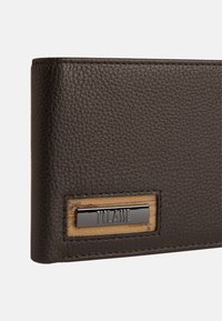 Portafoglio in pelle marrone con superficie texturizzata, etichetta rettangolare cucita con un dettaglio metallico e sfondo decorativo a motivo.