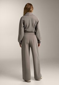 Šedý dvoudílný outfit se skládá z přiléhavého saka s vysokým límcem a širokými nohavicemi, vyrobený z měkké texturované látky s hladkým povrchem.