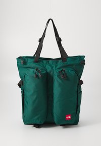 Saco de nylon verde com dois bolsos frontais com fecho, alças pretas ajustáveis e um pequeno logo vermelho na parte inferior direita.