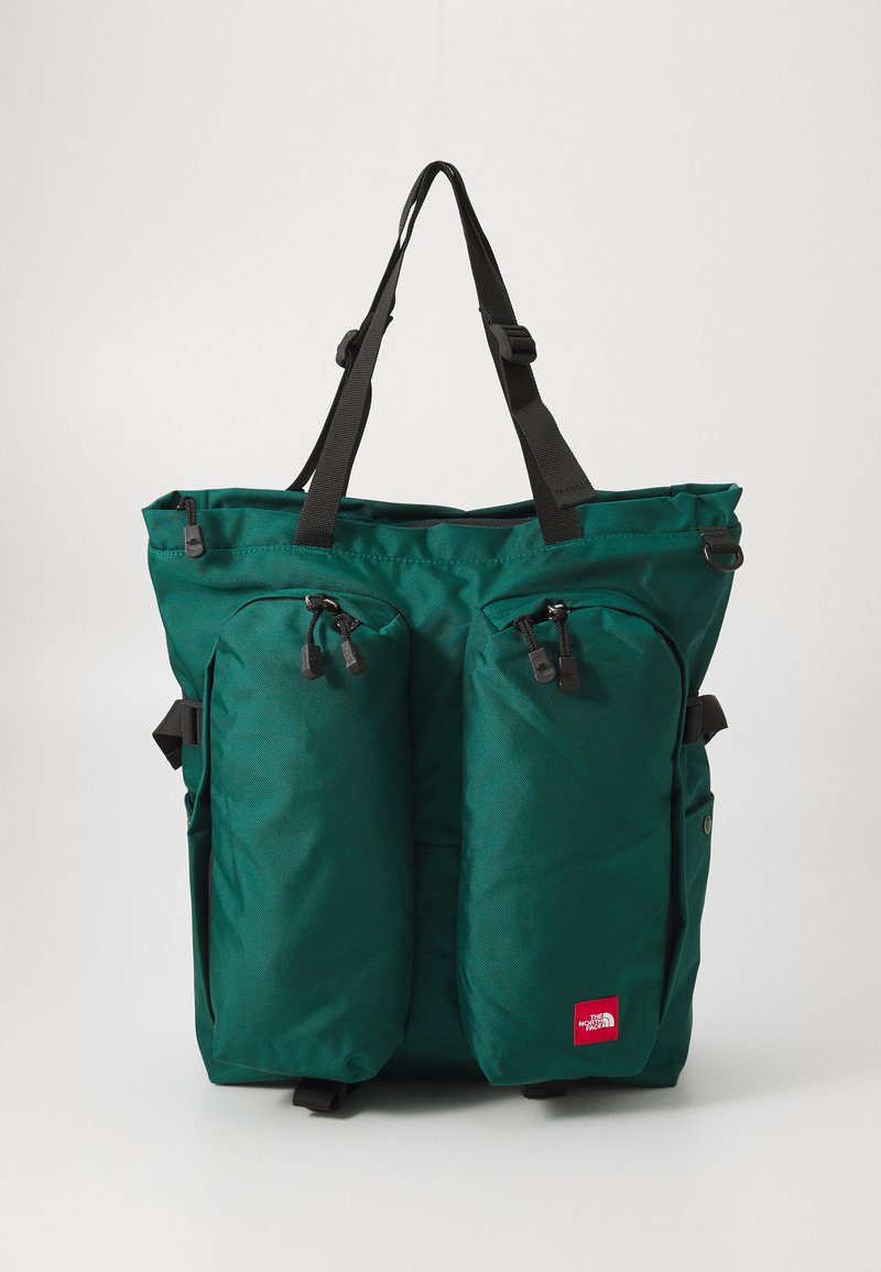 Saco de nylon verde com dois bolsos frontais com fecho, alças pretas ajustáveis e um pequeno logo vermelho na parte inferior direita.