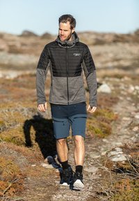 Giacca isolante nera e grigia con cappuccio, abbinata a pantaloni corti blu scuro con tasche, indossata con calzini neri e scarpe da ginnastica.